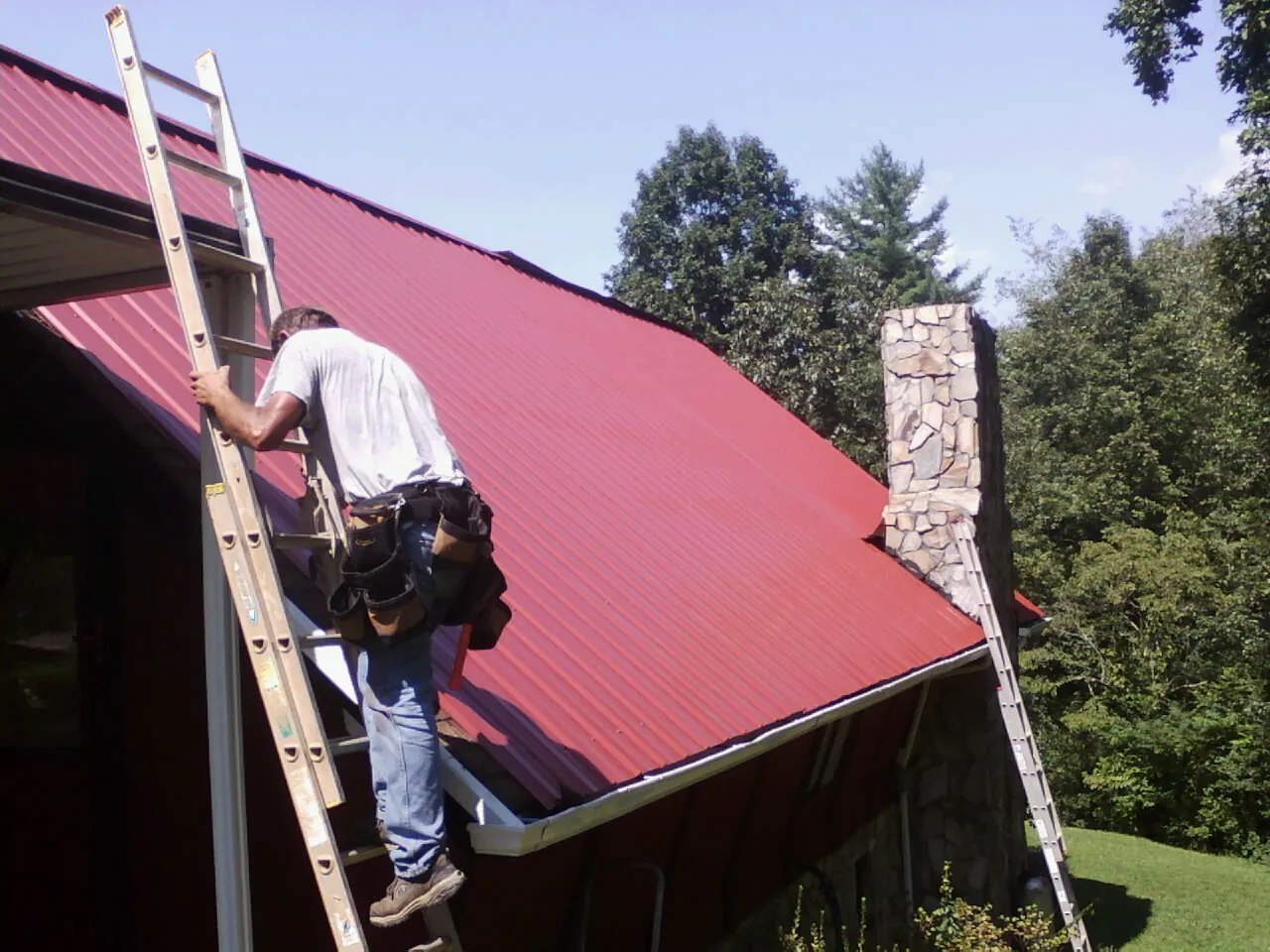 Roofer on a ladder beside a red metal roof and stone chimney — Soffit & Fascia Repair by Roofing Brothers in Hunters